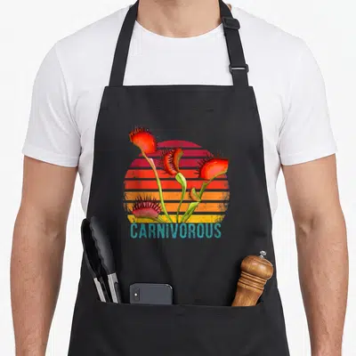 Tablier de cuisine noir avec illustration de plantes carnivores rouges et d'un soleil couchant strié orange et rose, texte CARNIVOROUS en bas.