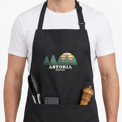 Tablier de cuisine noir avec illustration de soleil et arbres verts mentionnant Astoria Oregon