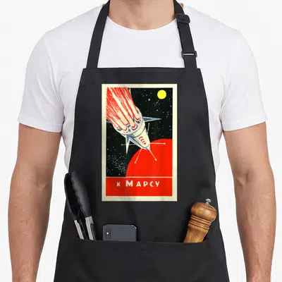 Tablier de cuisine noir avec illustration retro d'une fusée rouge et blanche vers une planète rouge et l'inscription K Mars en bas.