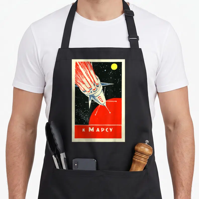 Tablier de cuisine noir avec illustration retro d'une fusée rouge et blanche vers une planète rouge et l'inscription K Mars en bas.