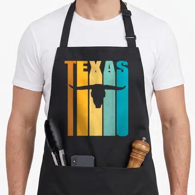 Tablier de cuisine noir avec linscription TEXAS en bandes de couleur orange jaune et turquoise surplombant une tête de vache Longhorn grise.
