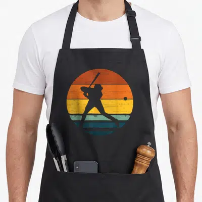 Tablier de cuisine noir avec silhouette de softballeuse sur fond de soleil couchant aux bandes orange jaune et bleu.