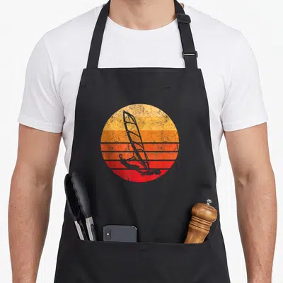 Tablier de cuisine noir avec illustration circulaire d'une planche à voile sur un soleil couchant aux bandes orange et rouges.