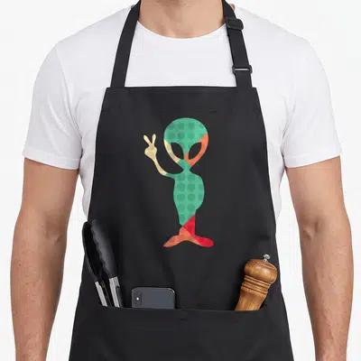 Tablier de cuisine noir avec un alien vert à pois faisant un signe de la paix dans un style rétro coloré.