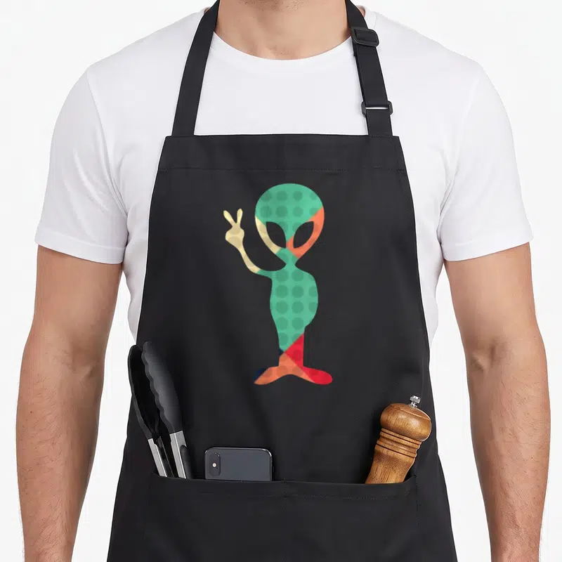 Tablier de cuisine noir avec un alien vert à pois faisant un signe de la paix dans un style rétro coloré.