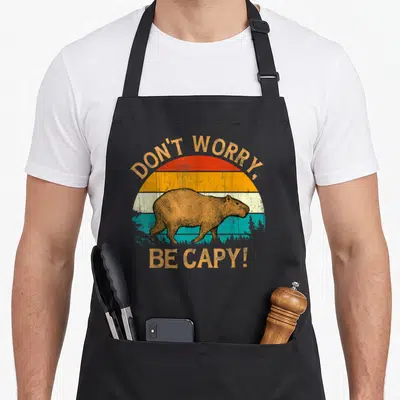 Tablier de cuisine noir avec illustration d'un capybara et texte Don't Worry Be Capy sur fond de soleil coloré.