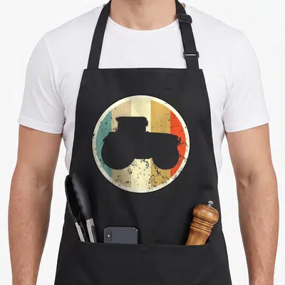 Tablier de cuisine noir avec illustration circulaire d'un tracteur gris sur bandes de couleurs pastel et effet vieilli.
