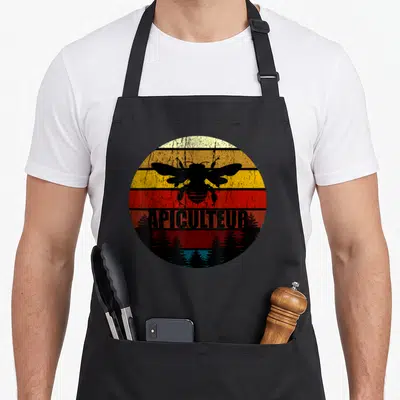 Tablier de cuisine noir avec motif circulaire rétro montrant une abeille et linscription apiculteur au-dessus de silhouettes darbres.