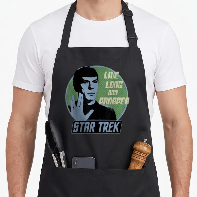 Tablier de cuisine noir avec illustration circulaire de Spock et texte Live Long and Prosper en lettres jaunes sur fond vert rayé.