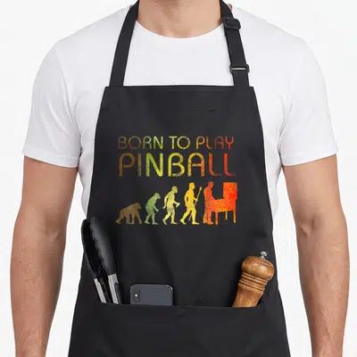 Tablier de cuisine noir avec illustration évolutive Born to Play Pinball en dégradé de couleurs vintage.