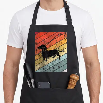 Tablier de cuisine noir avec illustration d'un teckel noir sur fond de bandes diagonales colorées et effet vieilli.