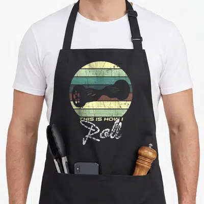 Tablier de cuisine noir avec illustration d'hoverboard vintage, cercle aux bandes colorées et texte This Is How I Roll en typographie décontractée.