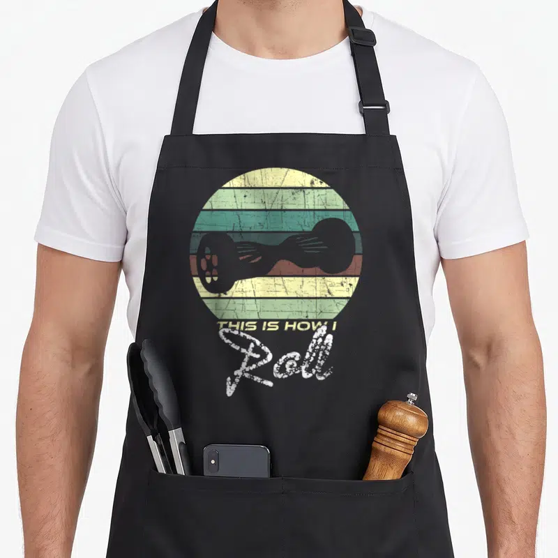 Tablier de cuisine noir avec illustration d'hoverboard vintage, cercle aux bandes colorées et texte This Is How I Roll en typographie décontractée.