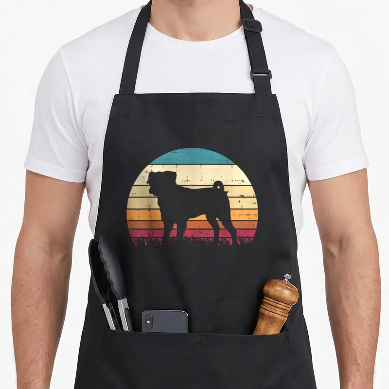 Tablier de cuisine noir avec silhouette de carlin devant un soleil couchant rétro multicolore et des touffes d'herbe stylisées.