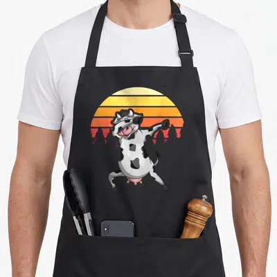 Tablier de cuisine noir avec une vache blanche à taches noires portant des lunettes de soleil et posant devant un soleil orange strié.