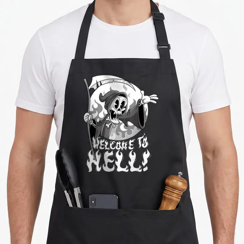 Tablier de cuisine noir avec un squelette cartoon tenant une faux et le texte Bienvenue en hell émergeant de flammes blanches.