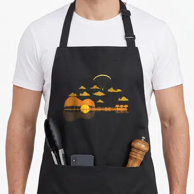 Tablier de cuisine noir avec illustration orange d'un parapente et d'un coucher de soleil formant une guitare dans l'eau.
