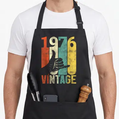 Tablier de cuisine noir avec l'année 1976, un signe Shaka et le mot Vintage en lettres colorées.