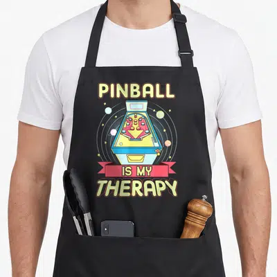 Tablier de cuisine noir avec illustration de flipper spatial et texte Pinball is my therapy en lettres jaunes et roses.