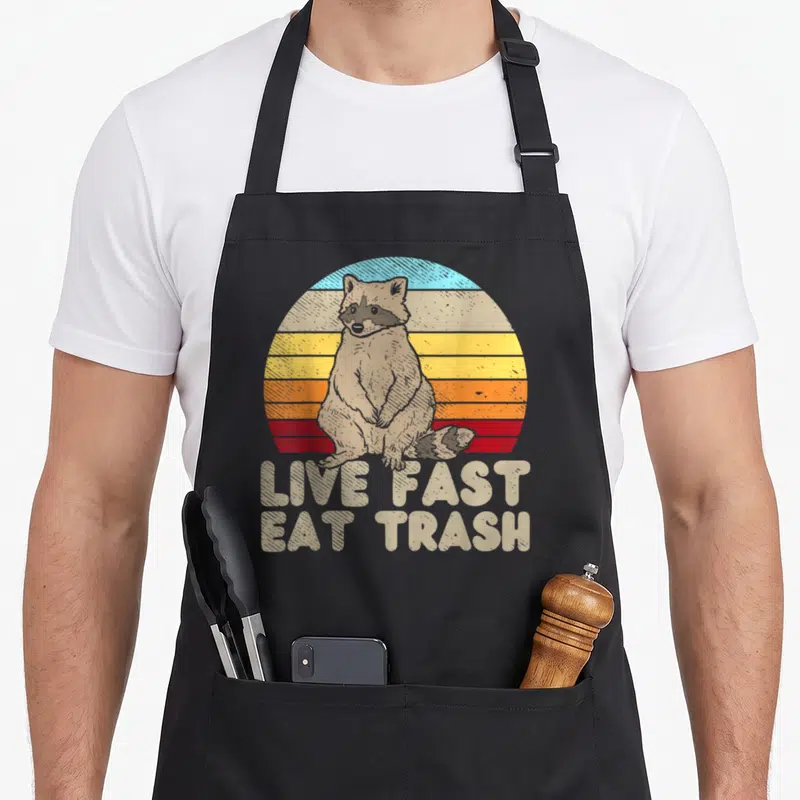 Tablier de cuisine noir avec illustration d'un raton laveur devant un soleil couchant retro et le texte Live Fast Eat Trash en bas.