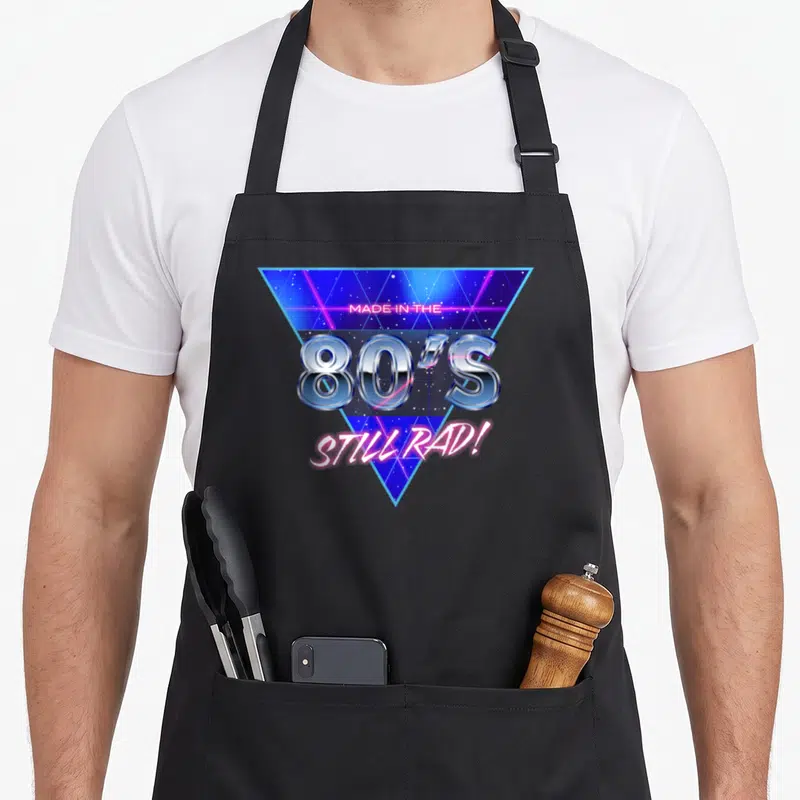 Tablier de cuisine noir avec logo triangulaire bleu néon et texte chromé 80's Still Rad sur fond étoilé.