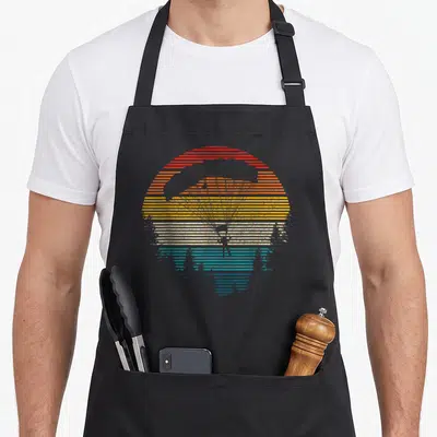 Tablier de cuisine noir avec illustration rétro d'un parachutiste sur fond de soleil rayé multicolore.