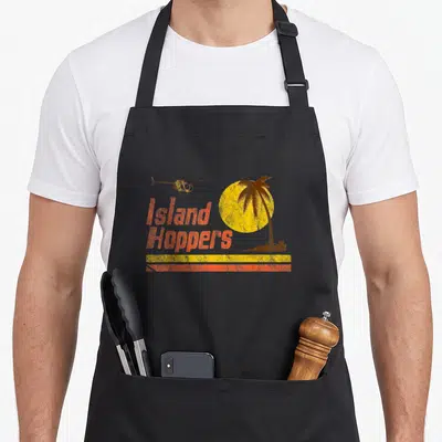 Tablier de cuisine noir avec illustration Island Hoppers, soleil jaune, palmier et hélicoptère orange style rétro.