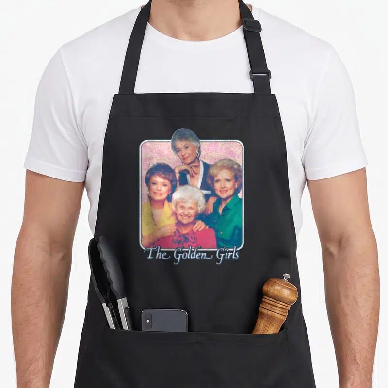Tablier de cuisine noir avec une illustration groupée des quatre personnages de The Golden Girls sur fond rose texturé.