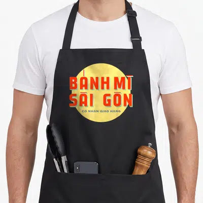 Tablier de cuisine noir avec motif circulaire jaune affichant Banh Mi Sai Gon en lettres rouges et un slogan en vietnamien.
