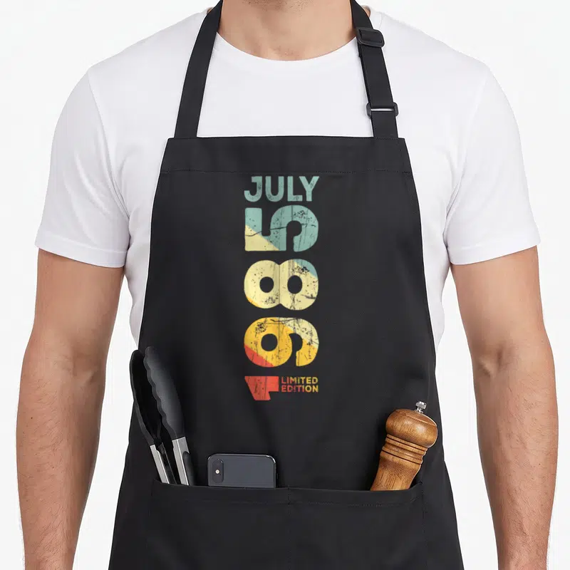 Tablier de cuisine noir avec typographie verticale colorée indiquant Juillet 1985 et Limited Edition sur fond gris uni.