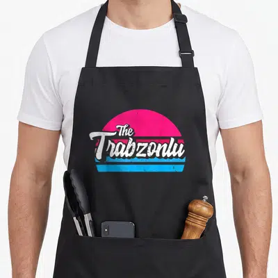 Tablier de cuisine noir avec le logo The Trabzonlu en blanc sur un soleil rose et des bandes bleues style rétro.