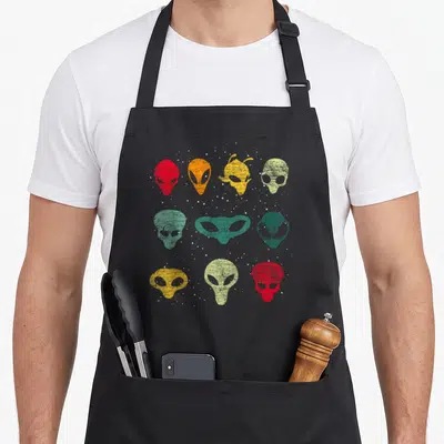 Tablier de cuisine noir avec neuf visages d'extraterrestres multicolores sur fond gris uni.