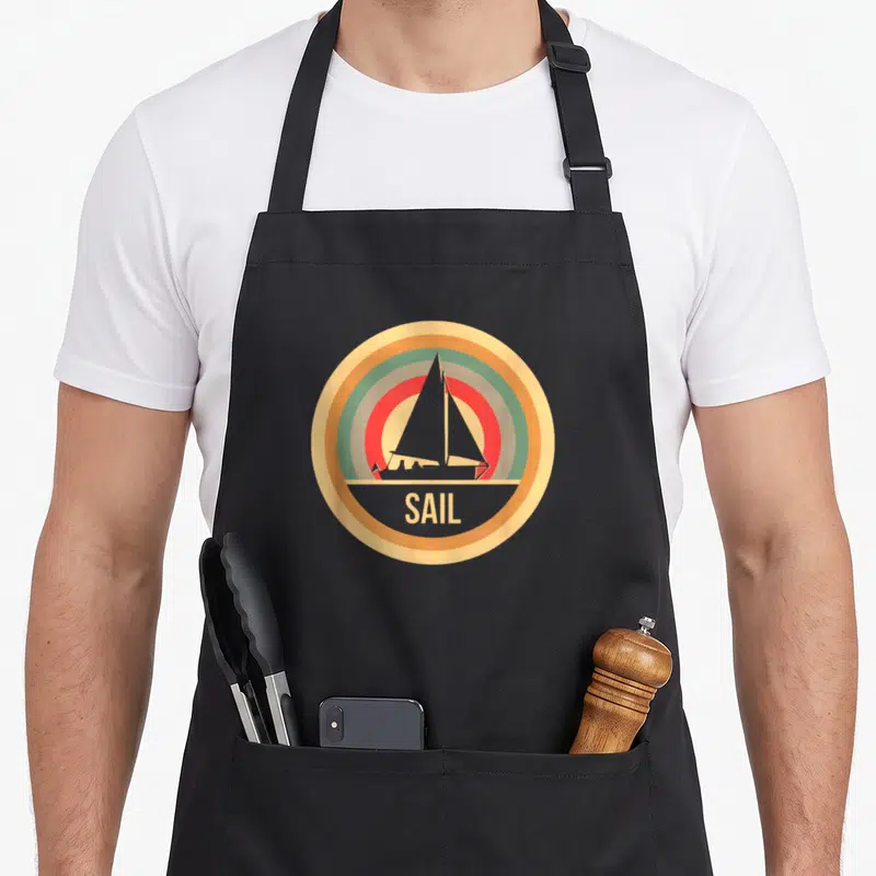 Tablier de cuisine noir avec logo circulaire rétro montrant un voilier et le mot SAIL en lettres capitales jaunes.