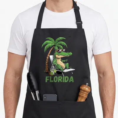 Tablier de cuisine noir avec illustration d'un crocodile aux lunettes de soleil sur un transat à côté d'un palmier et du mot Florida.
