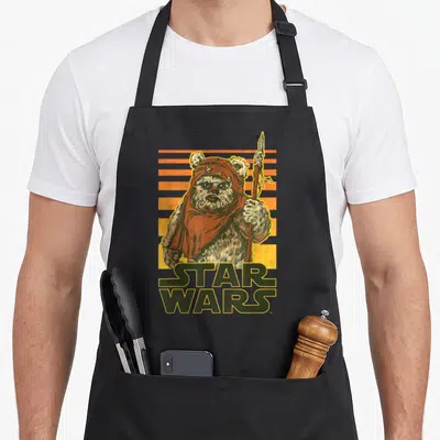 Tablier de cuisine noir avec illustration rétro de Wicket Ewok tenant une lance sous un soleil couchant orange.