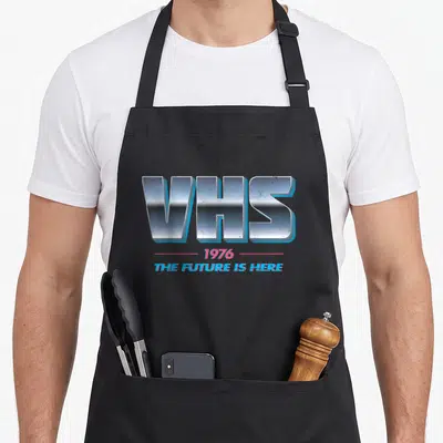 Tablier de cuisine noir avec le logo VHS 1976 The Future Is Here en lettres métalliques bleues et roses.