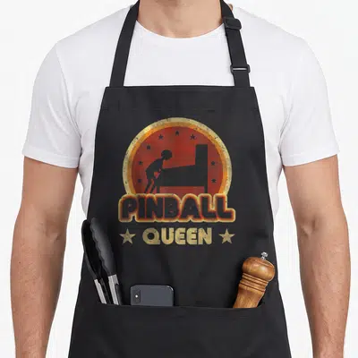 Tablier de cuisine noir avec logo Pinball Queen, silhouette joueur et fond rouge étoilé style rétro.
