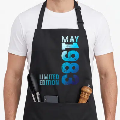 Tablier de cuisine noir avec typographie verticale bleue indiquant En mai 1983 Limited Edition sur fond uni.