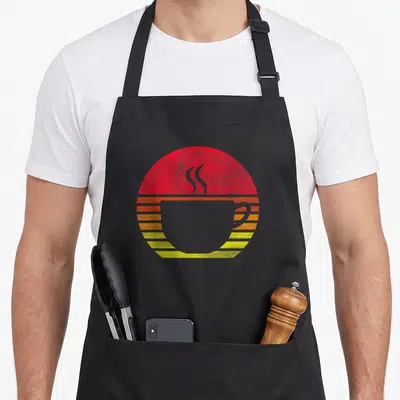 Tablier de cuisine noir avec une illustration circulaire de tasse de café sur fond de soleil couchant aux teintes rouges et jaunes.