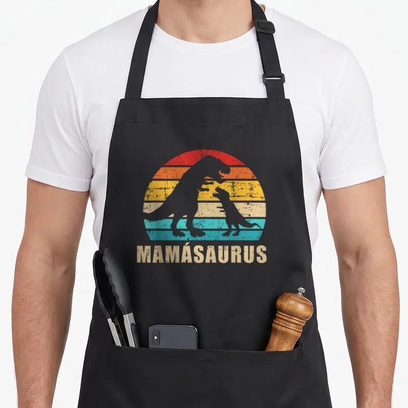 Tablier de cuisine noir avec silhouette de dinosaures et texte Mamásaurus sur fond coloré rétro.