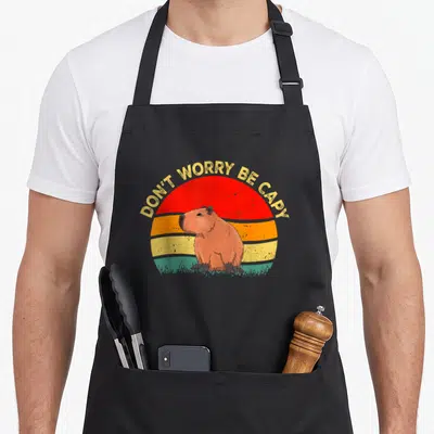Tablier de cuisine noir avec illustration de capybara et texte DON'T WORRY BE CAPY sur fond de soleil coloré.