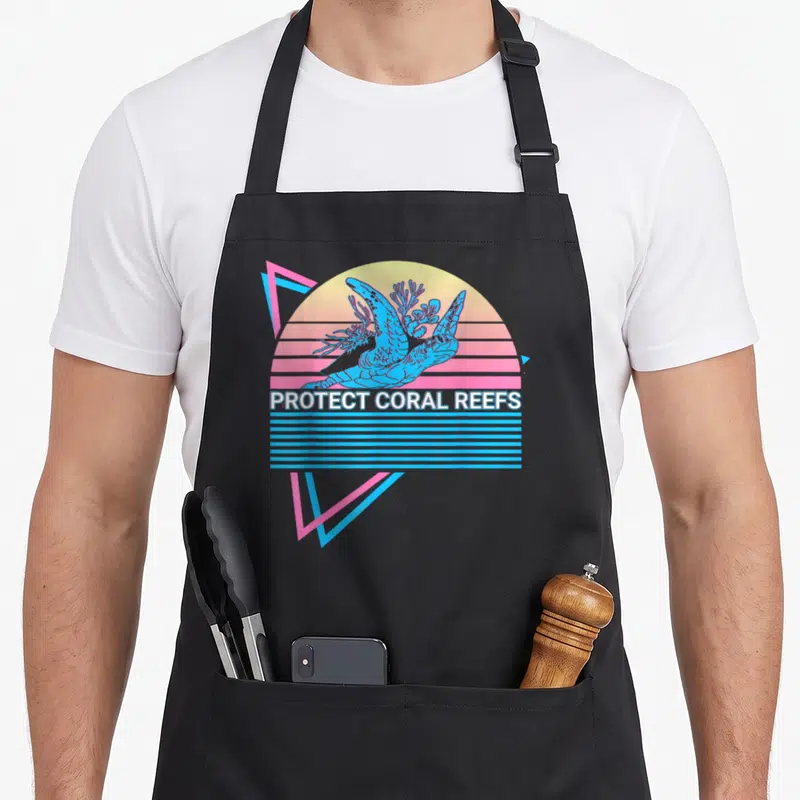 Tablier de cuisine noir avec illustration rétro d'une tortue et du texte Protect Coral Reefs sur fond de soleil couchant.