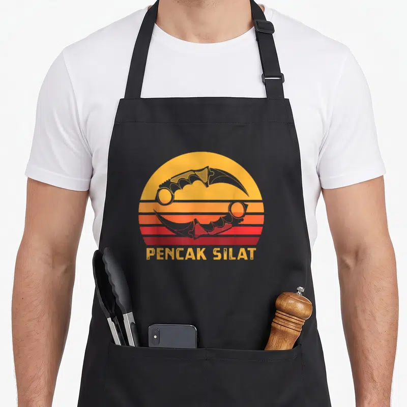 Tablier de cuisine noir avec illustration rétro de deux karambits et le texte Pencak Silat en jaune sur fond de soleil rayé orange et rouge.