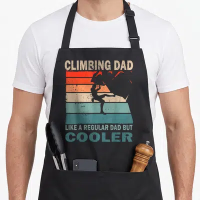 Tablier de cuisine noir avec design rétro Climbing Dad et silhouette de grimpeur sur fond de bandes colorées.