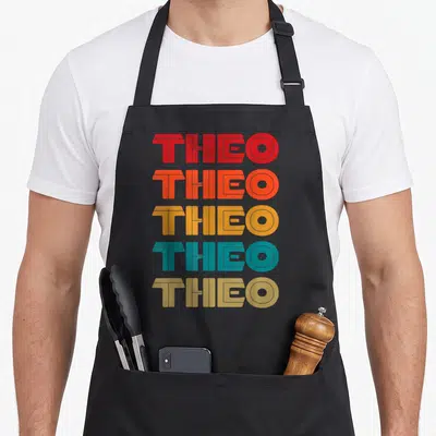 Tablier de cuisine noir avec le nom THEO répété en lettres colorées style années soixante-dix.