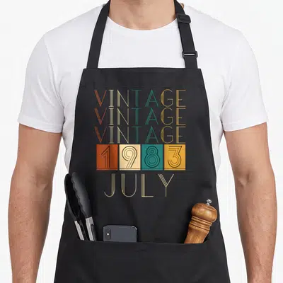 Tablier de cuisine noir avec typographie rétro Vintage 1983 et July en lettres fines multicolores.