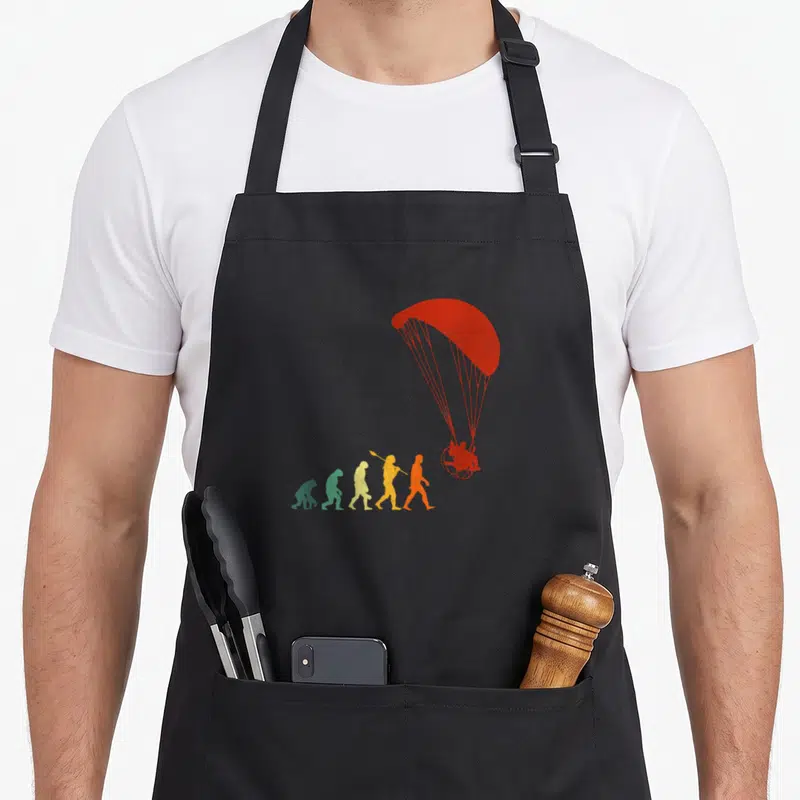 Tablier de cuisine noir avec une illustration colorée montrant l'évolution humaine vers un paramoteur rouge.
