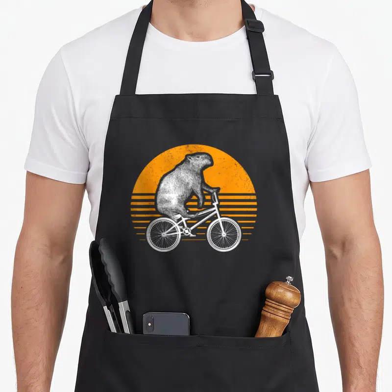 Tablier de cuisine noir avec illustration d'un capybara sur vélo rétro devant un soleil orange strié.
