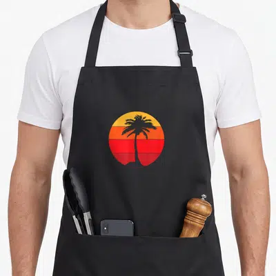 Tablier de cuisine noir avec illustration circulaire d'un palmier sur fond de soleil dégradé orange et rouge