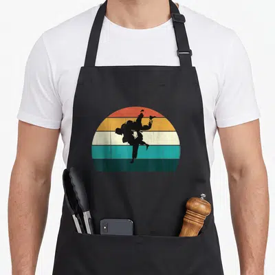 Tablier de cuisine noir avec une illustration rétro de deux lutteurs en silhouette noire effectuant un flip sur un demi-soleil multicolore à bandes horizontales.